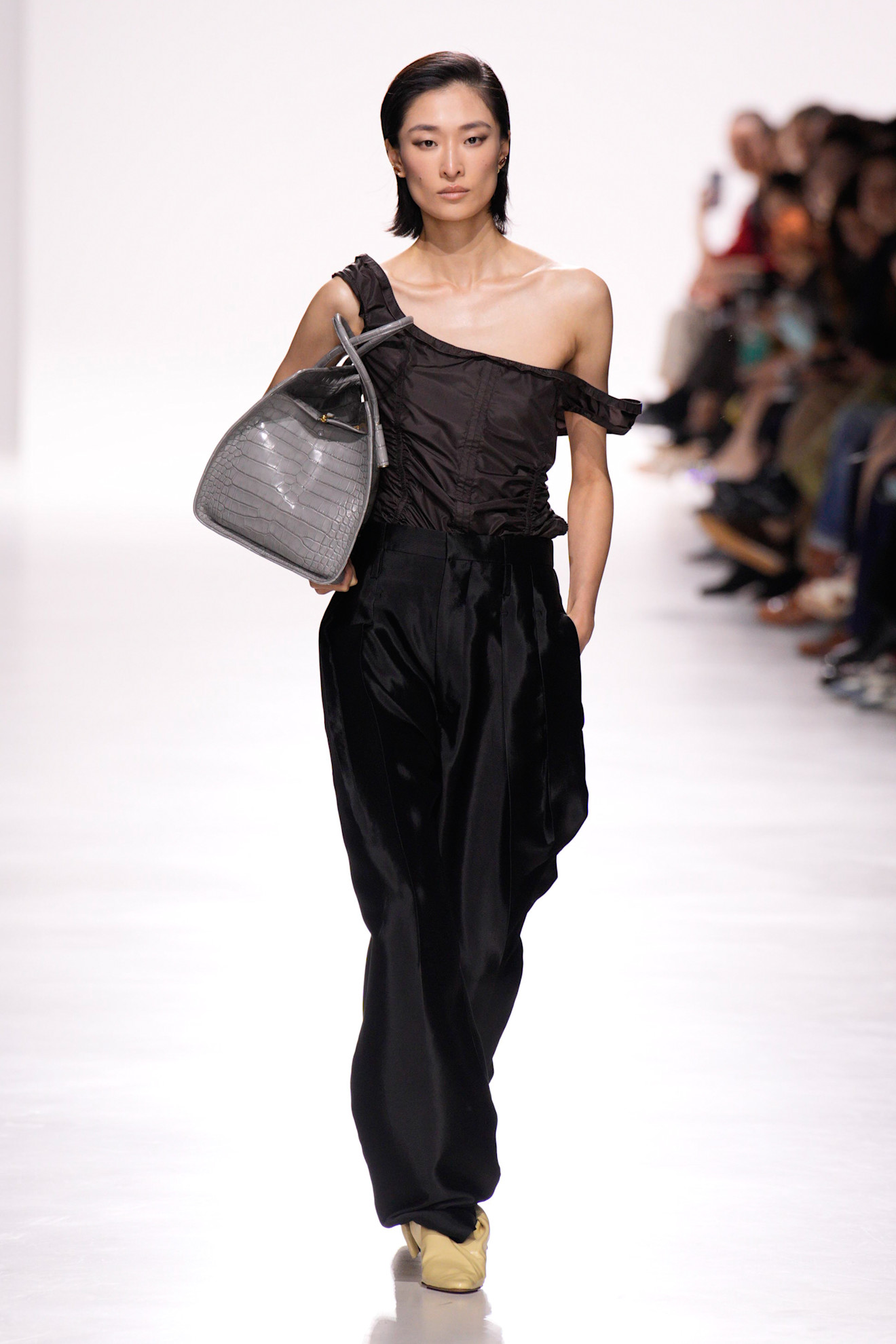 Bottega Veneta model walking in the S/S 26 show wearing a black off-the-shoulder top and carrying a gray croc tote bag.