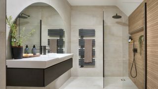 bathroom with walk in shower, grey floating sink vanity, circular mirror, cream tiles and grey towel radiator
