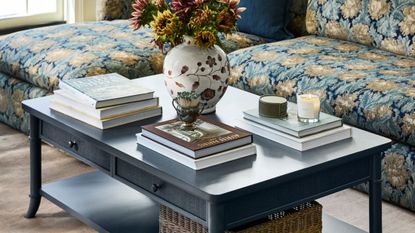 Two-tier navy blue coffee table with storage styled with stacks of coffee table books, a floral-patterned vase, match strike, and lit candles.