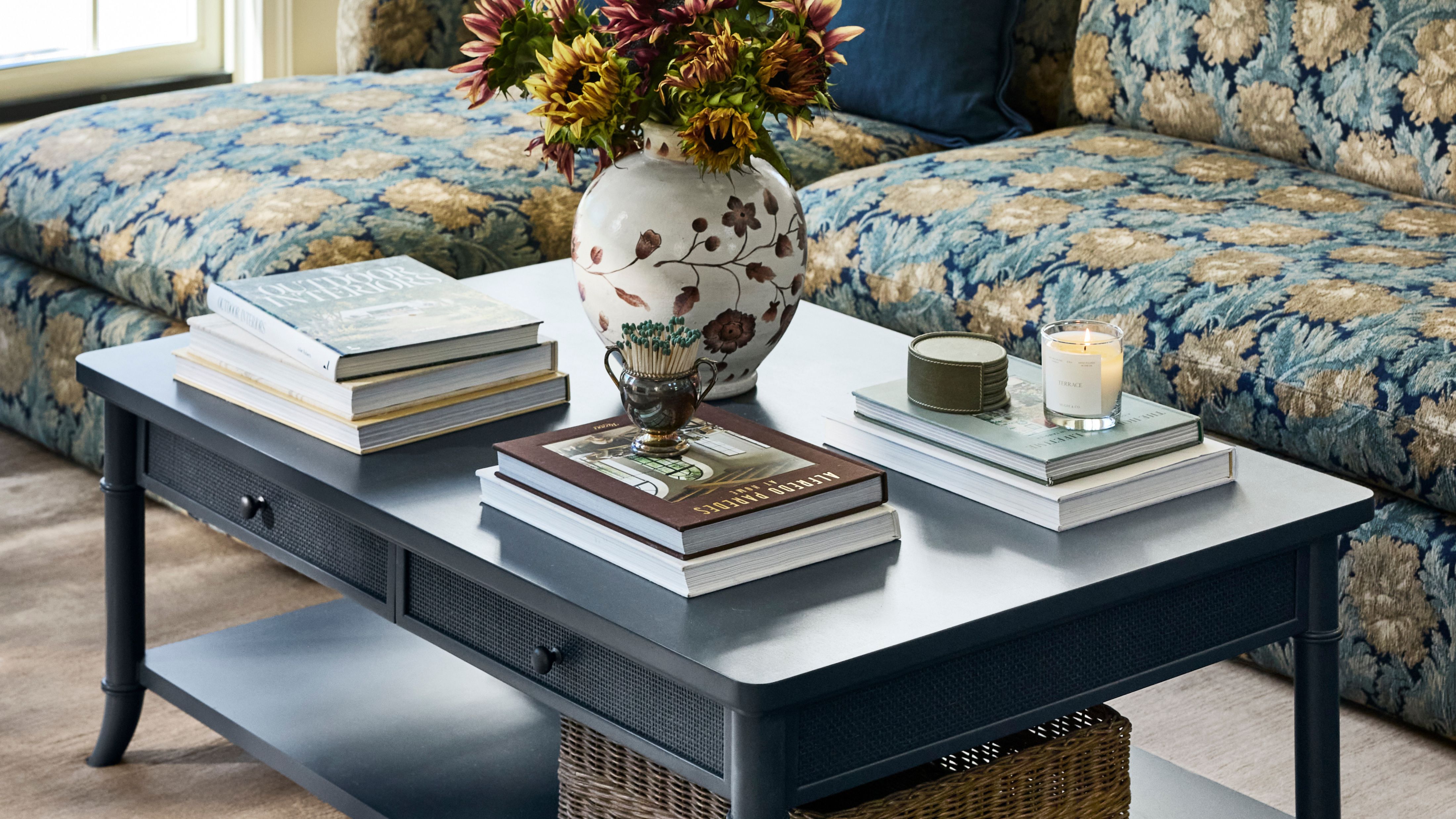 Two-tier navy blue coffee table with storage styled with stacks of coffee table books, a floral-patterned vase, match strike, and lit candles.