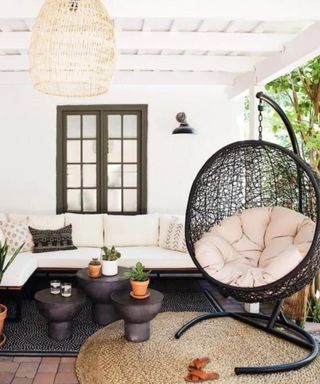 A backyard area with a white house, a rattan pendant light, a black egg chair, and an L-shaped corner seat