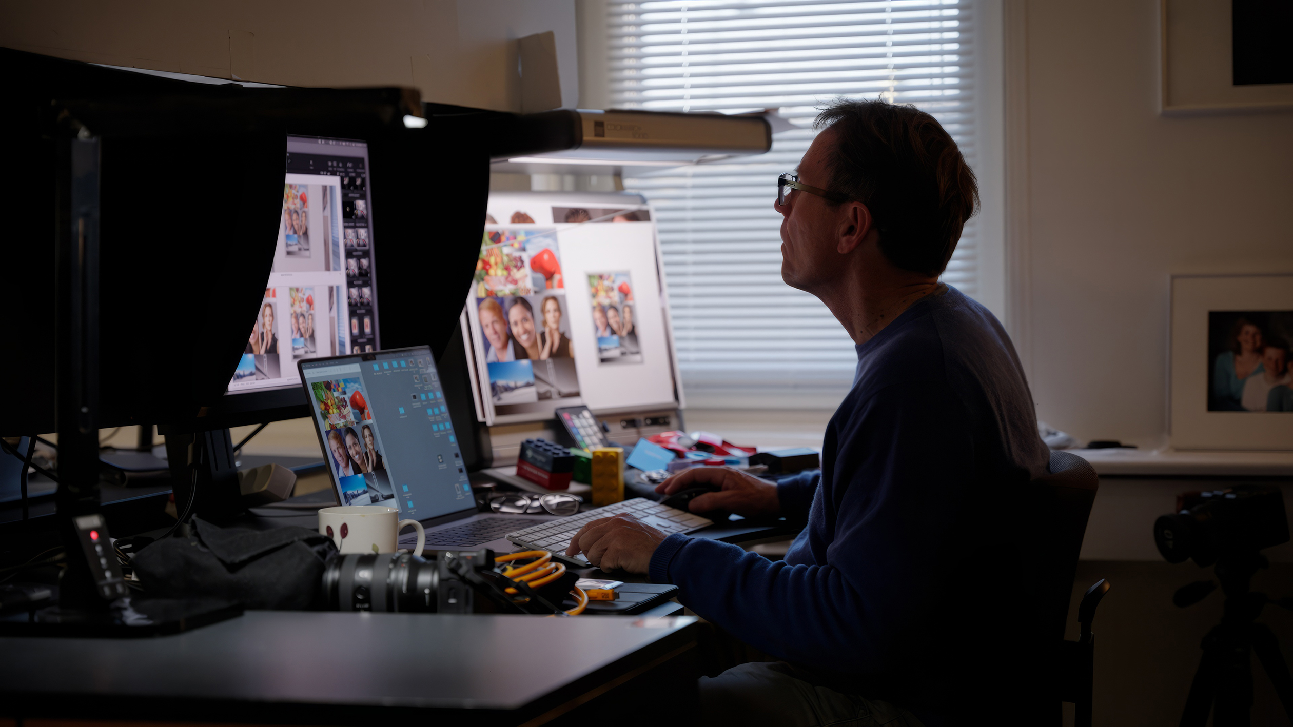 Man working on photo editing at a dual-monitor desk setup in a dimly lit room with photography equipment nearby