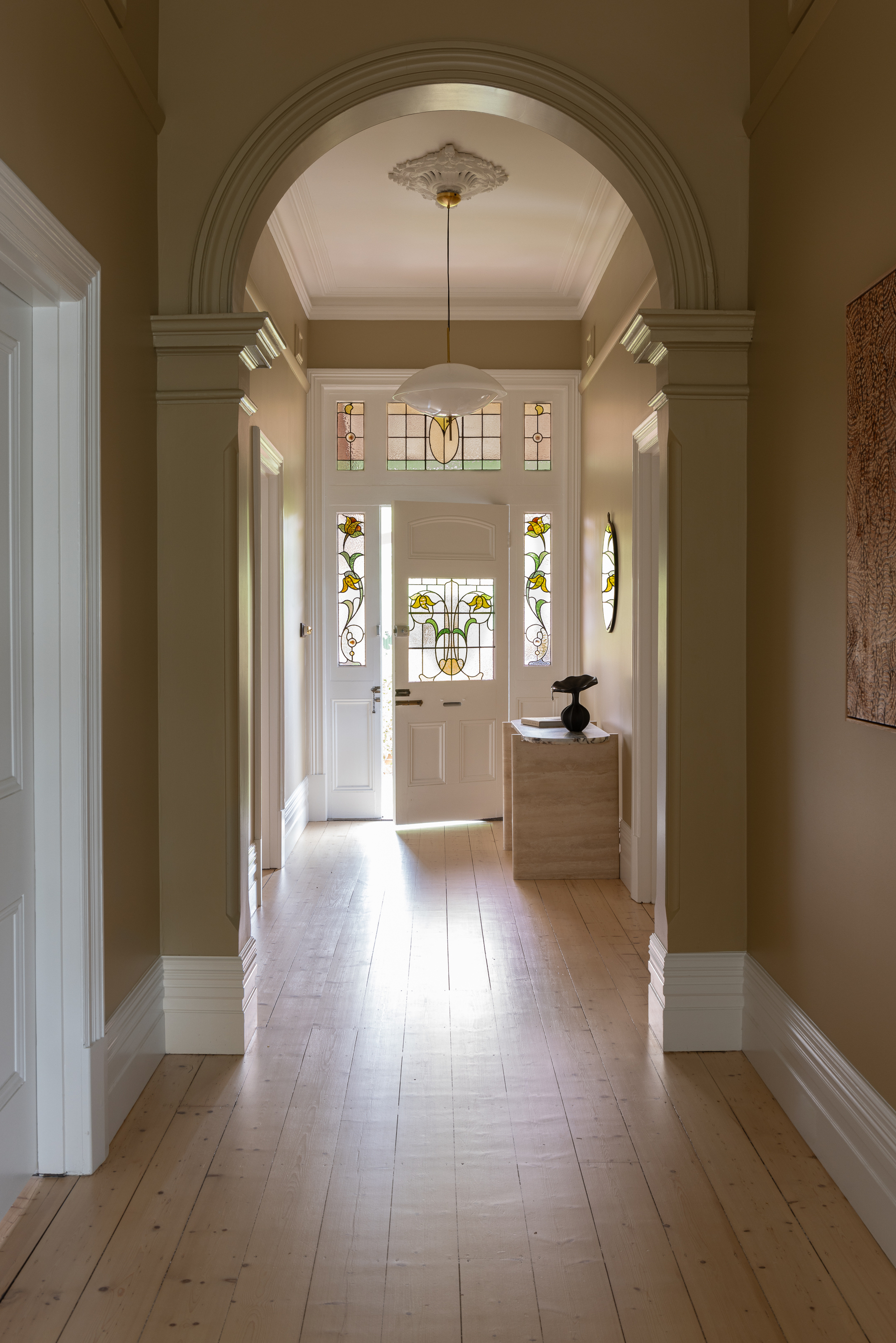 hallway of historic home with arch, caramel paint on walls with white trim, pine floorboards, front door with stained glass, and a modern console table styled with a lamp