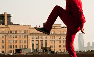 Close up view of a dancer- Red suit, bent raised knee, building behind