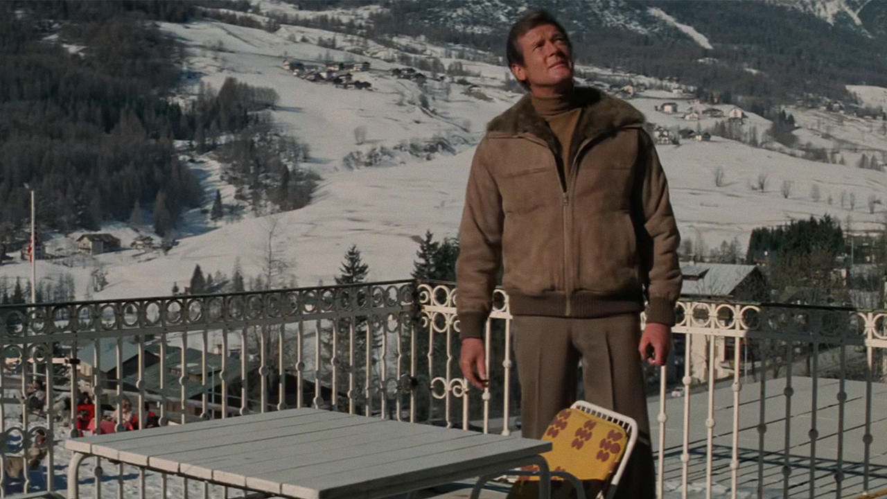 Roger Moore in a brown coat standing on a balcony over a ski town in For Your Eyes Only