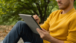 Amazon Kindle Scribe in a case held in hand by a person sitting while leaning against a tree outdoors