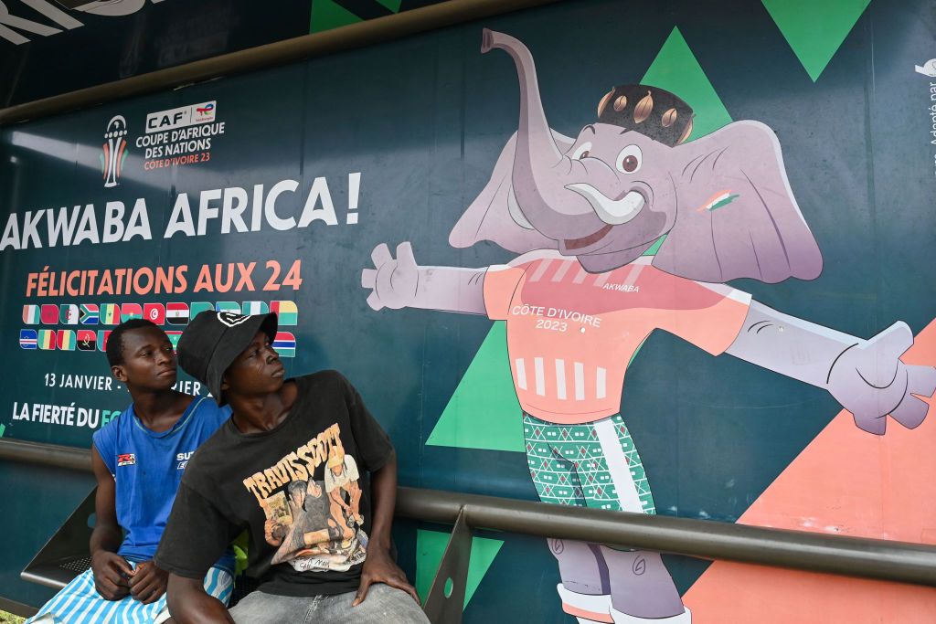 People sit at a bus stop decorated with the 2024 Africa Cup of Nations (CAN) official mascot &#039;Awkwaba&#039; in Abidjan on January 8, 2024. Ivory Coast is getting ready to host the 34th Africa Cup of Nations (CAN), which begins on January 13, 2024.