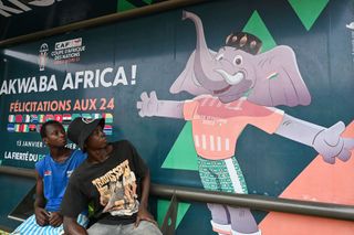 People sit at a bus stop decorated with the 2024 Africa Cup of Nations (CAN) official mascot 'Awkwaba' in Abidjan on January 8, 2024. Ivory Coast is getting ready to host the 34th Africa Cup of Nations (CAN), which begins on January 13, 2024.