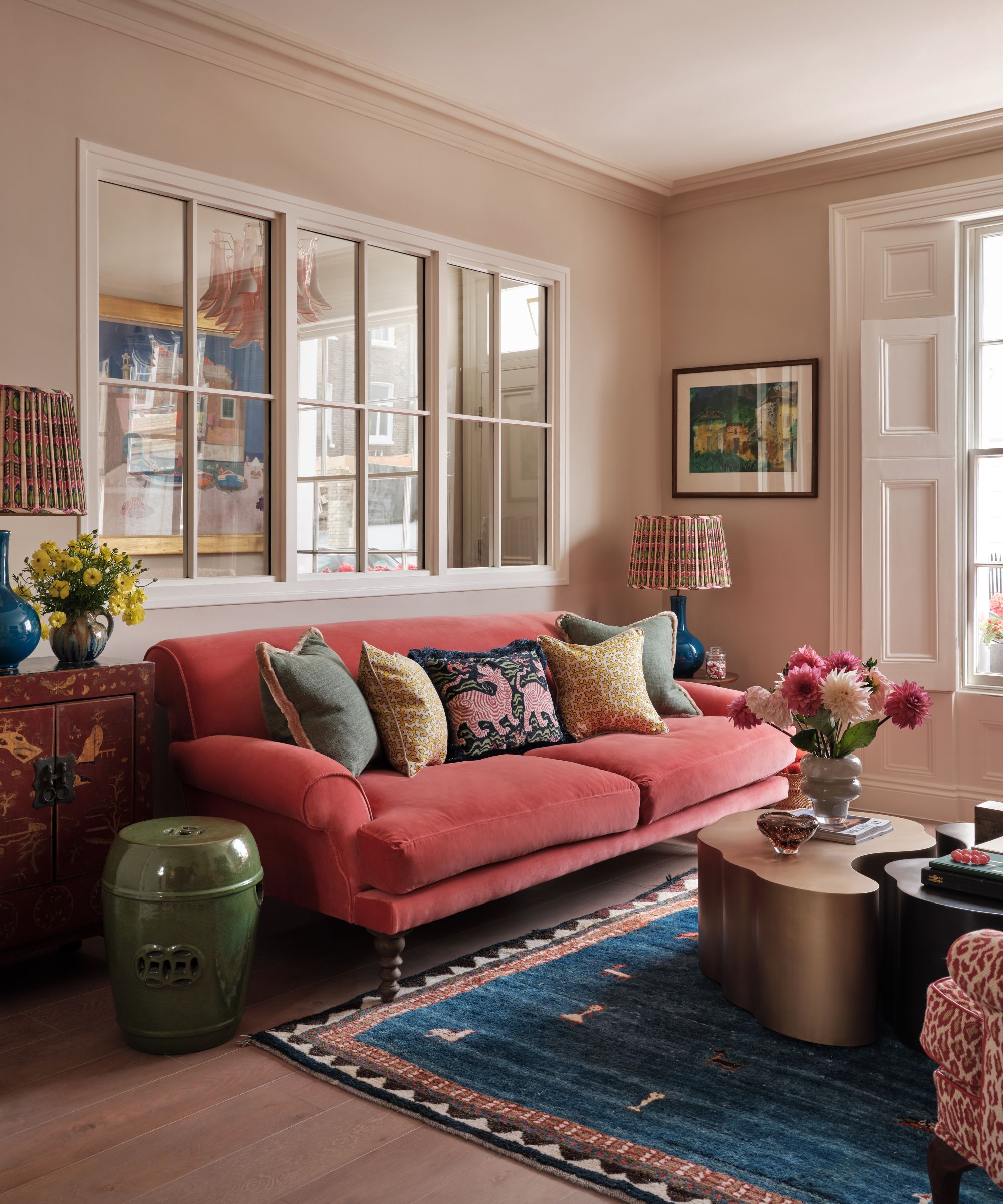 A layered, colorful living room with a pink couch, assorted cushions, a large blue rug, and abstract gold coffee table with a large vase of pink flowers. The walls are a neutral peach, and to the right is a large window with white shutters. Above the couch is a large partition window. To either side of the couch are matching blue lamps with colorful lampshades, and to the left, it's on a brown cabinet alongside a vase of yellow flowers, with a green ceramic stool in front.