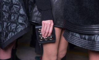 Close up shot of a model holding an accessory from the Diesel Black Gold collection. It is a square black leather accessory with round metallic embellishments. You can also see a close up view of some of the pieces in the collection made from leather and quilted fabrics