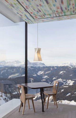 Interior view of AlpiNN restaurant featuring a table, two chairs, a low hanging asymmetric pendant light and wooden floors. The ceiling is multicoloured and views of the mountains can be seen through the floor to ceiling windows
