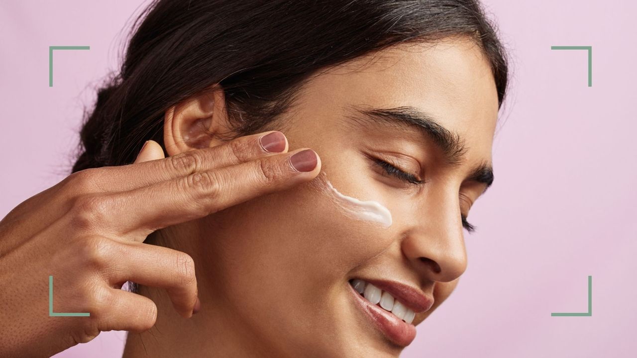 A woman applying retinol eye cream with her fingers on a pink background