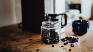 coffee beans spilling out of a pot with a coffee mug and coffee maker in the background