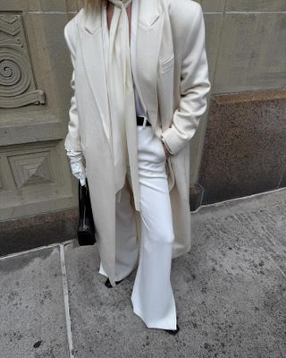 Christie wears an ivory coat, ivory blouse white trousers, black belt, and black heels.