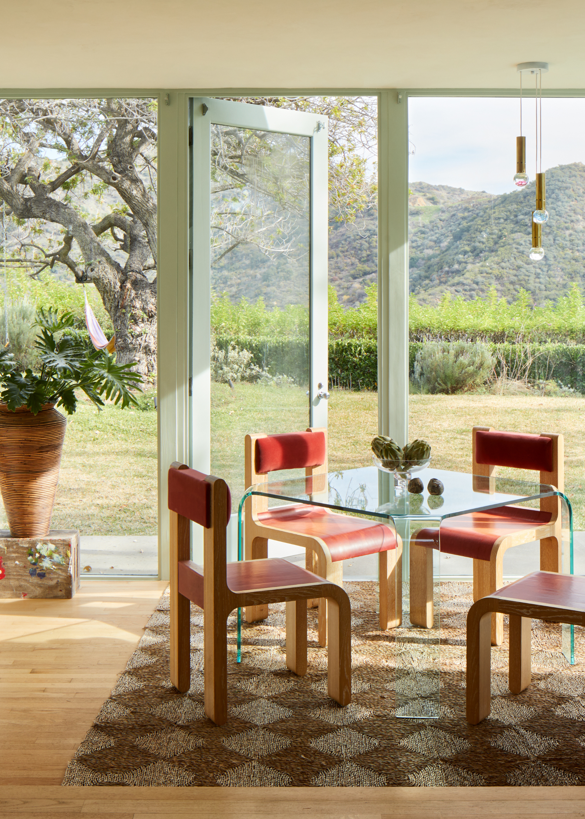 A dining room with a clear dining table, a harlequin rug, a set of red and wood chairs, beside a pedestal with a vase of stems, by a door leading to a garden