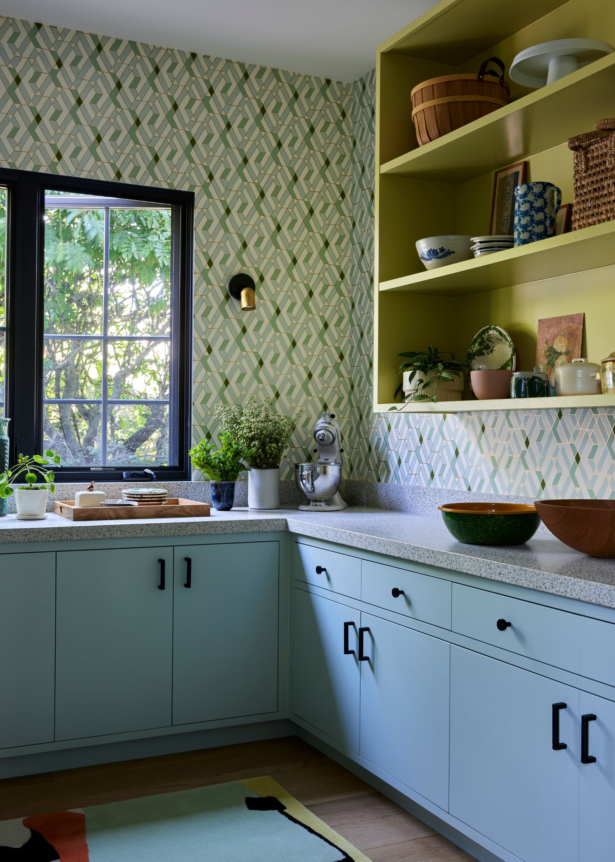 A pantry room with light blue lower kitchen cabinets, wallpapered walls, and a chartreuse wall-mounted open shelving unit with decorative objects on display.