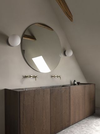 A minimalist bathroom with a wooden double vanity, wall mounted taps and a round mirror