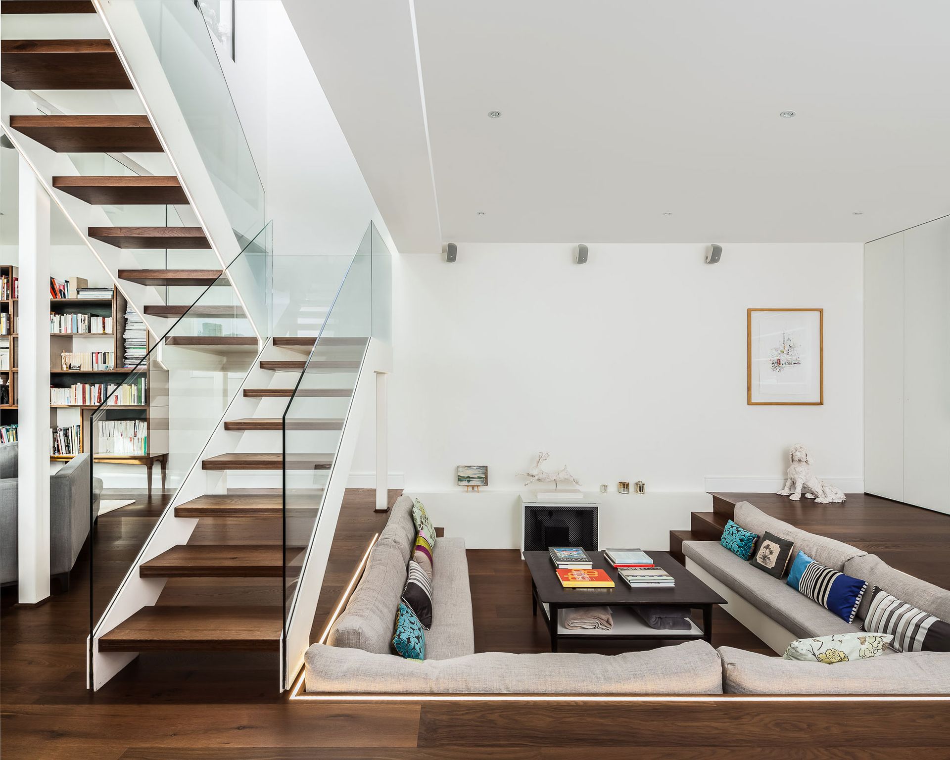 A living room space with sofas and a coffee table sunken into the floor next to a contemporary glass and wood staircase