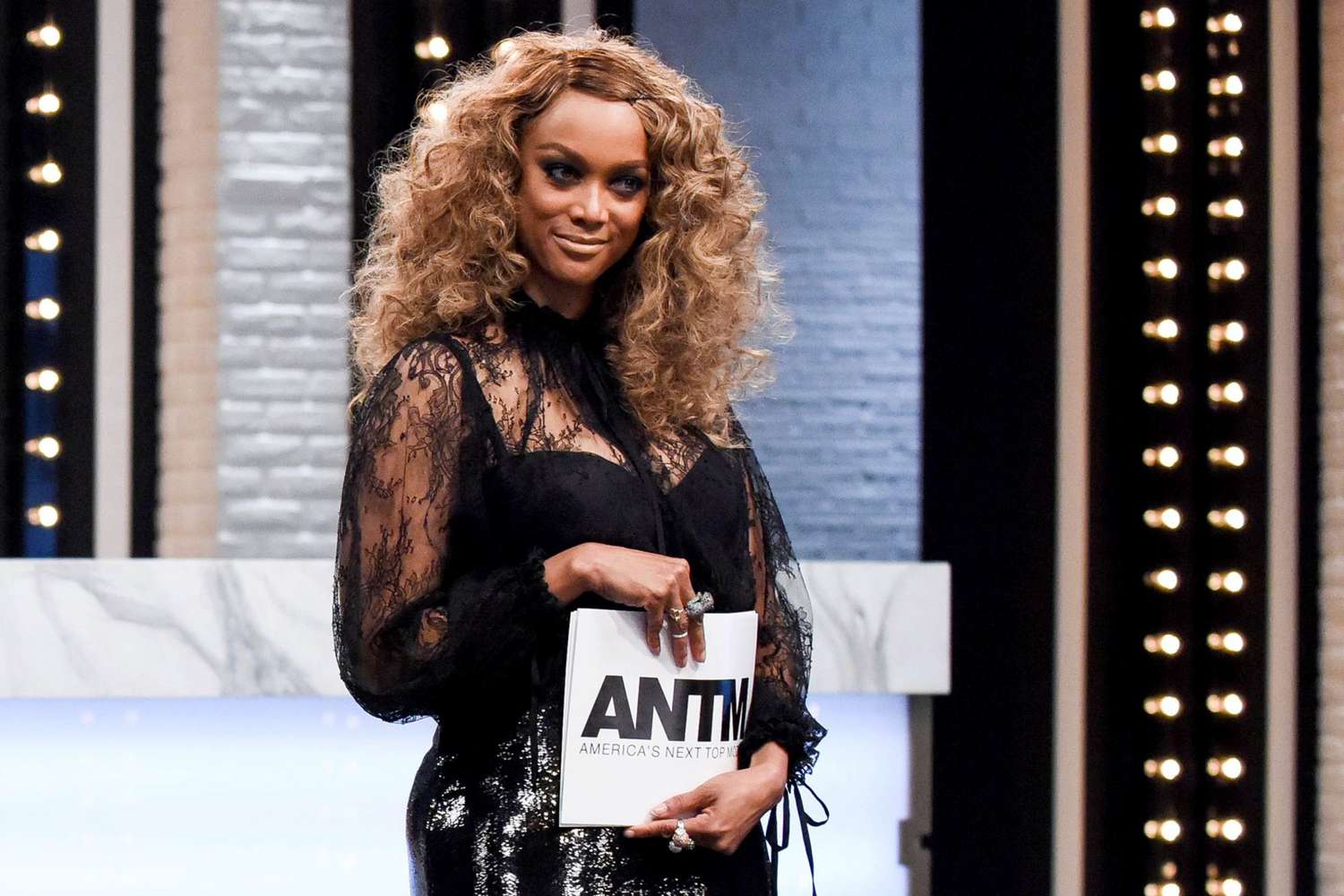 tyra banks wears a black lace dress and holds up photos while judging america&#039;s next top model