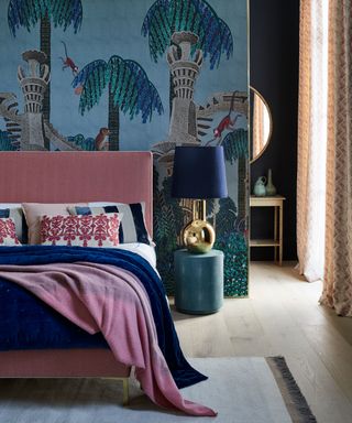 A luxury bedroom with a blue and green jungle tree print wall mural, pink upholstered bed and blue, pink and white accessories.