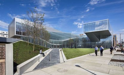Exterior view with a path that goes through a manicured green lawn to the building steel and glass panels. Photographed during the day 