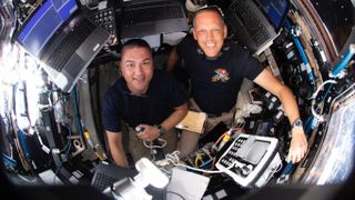 an overhead shot of two astronauts smiling in a small windowed room of the international space station, surrounded by computers and a window