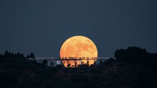 a large moon shines in the distance, in front is a long bridge-type observation deck with people standing on it. Their silhouettes' appears as a stark contrast on the lunar surface.