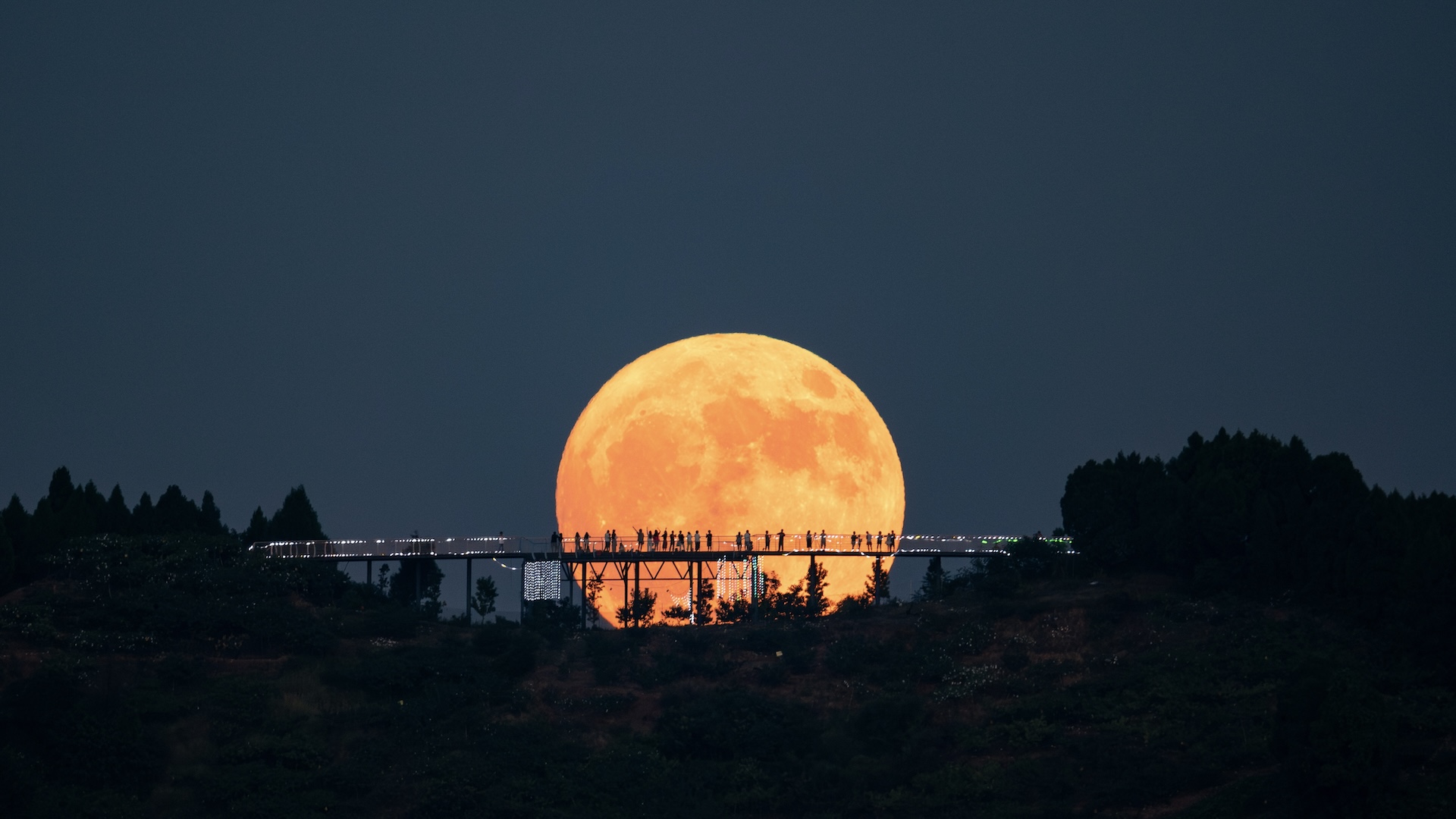 A large moon shines in the distance, in front of which is a long, bridge-like observation deck on which people stand. Their silhouettes form a strong contrast to the lunar surface.