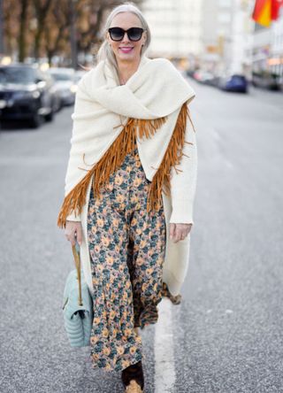 Best Ager Model and Influencer Petra van Bremen wearing a cream colored knitted coat by Antonia Zander, a cream colored scarf with brown leather fringe detail by Antonia Zander, a multicolored maxi dress with floral print by Ulla Johnson, sunglasses by Spektre Eyewear and brown vintage cowboy boots and a pastel mint green bag by Saint Laurent during a street style shooting on March 7, 2022