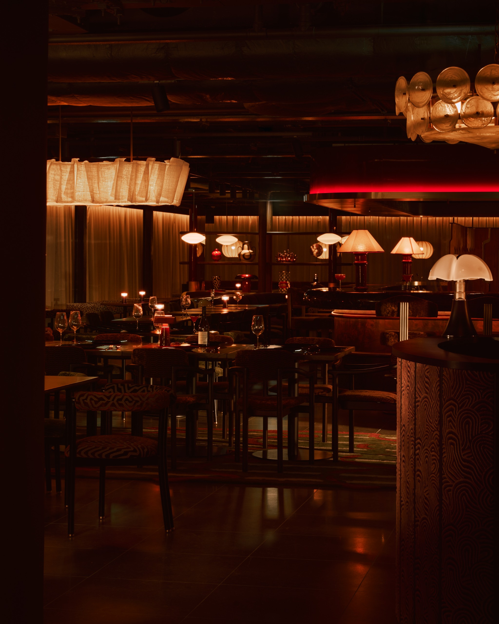 A red-tinted cocktail bar with golden-y pendant lights and table lamps, mid-century decor, a wooden floor decorated with a patterned geometric rug, and wrap-around beige curtains.