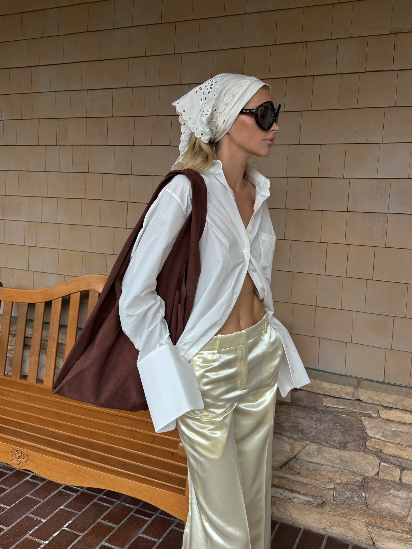 An image of a model wearing a white embroidered bandana, white button-down shirt, satin yellow trousers and a brown shoulder tote