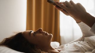 A woman lays on her back in bed with her smartphone held in front of her face with two hands after waking up from a night's sleep