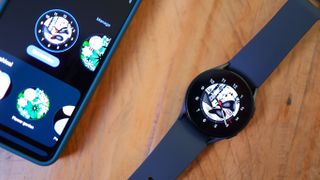 Bästa smartklocka: En Samsung Galaxy Watch 5 med ett mörkblått armband ligger på ett träbord bredvid en mobil med klockappen aktiv.