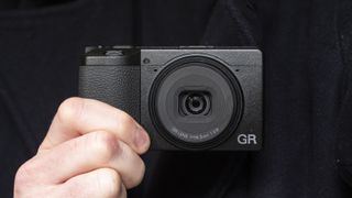 Ricoh GR IV compact camera in user's hand, with navy coat backdrop