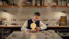 Designer Thomas Heatherwick sits at a table in a studio space examining an orb-shaped lamp