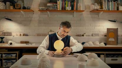 Designer Thomas Heatherwick sits at a table in a studio space examining an orb-shaped lamp
