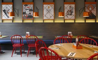 A restaurant with wooden tables, red chairs, wall paintings, orange wall lights and a blue sitting bench against the wall.