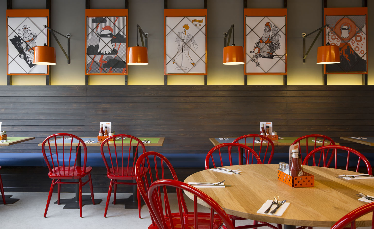 A restaurant with wooden tables, red chairs, wall paintings, orange wall lights and a blue sitting bench against the wall.