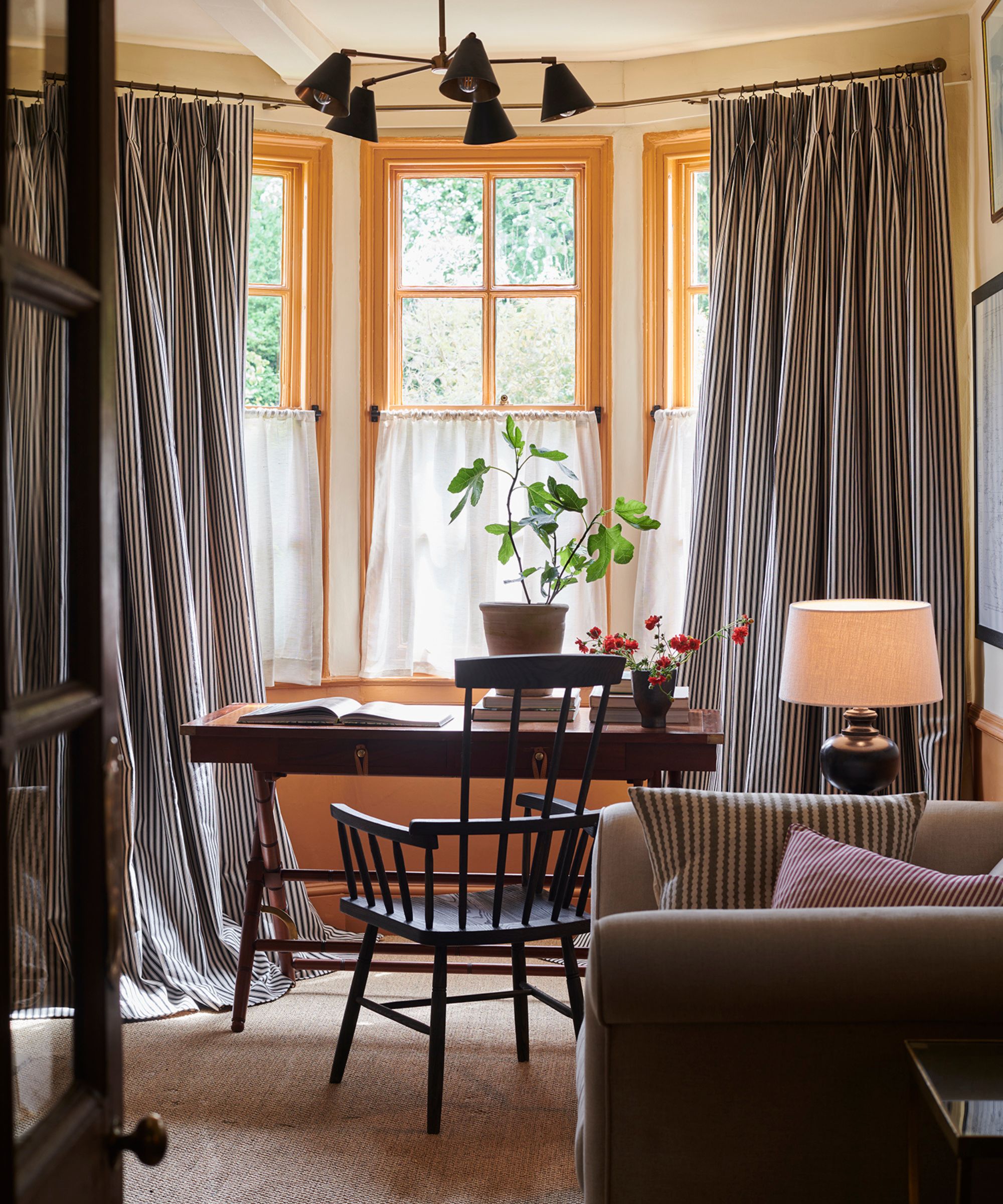 home office with a bay window painted yellow with striped tall curtains and white linen cage curtains with vintage office furniture and an arm chair