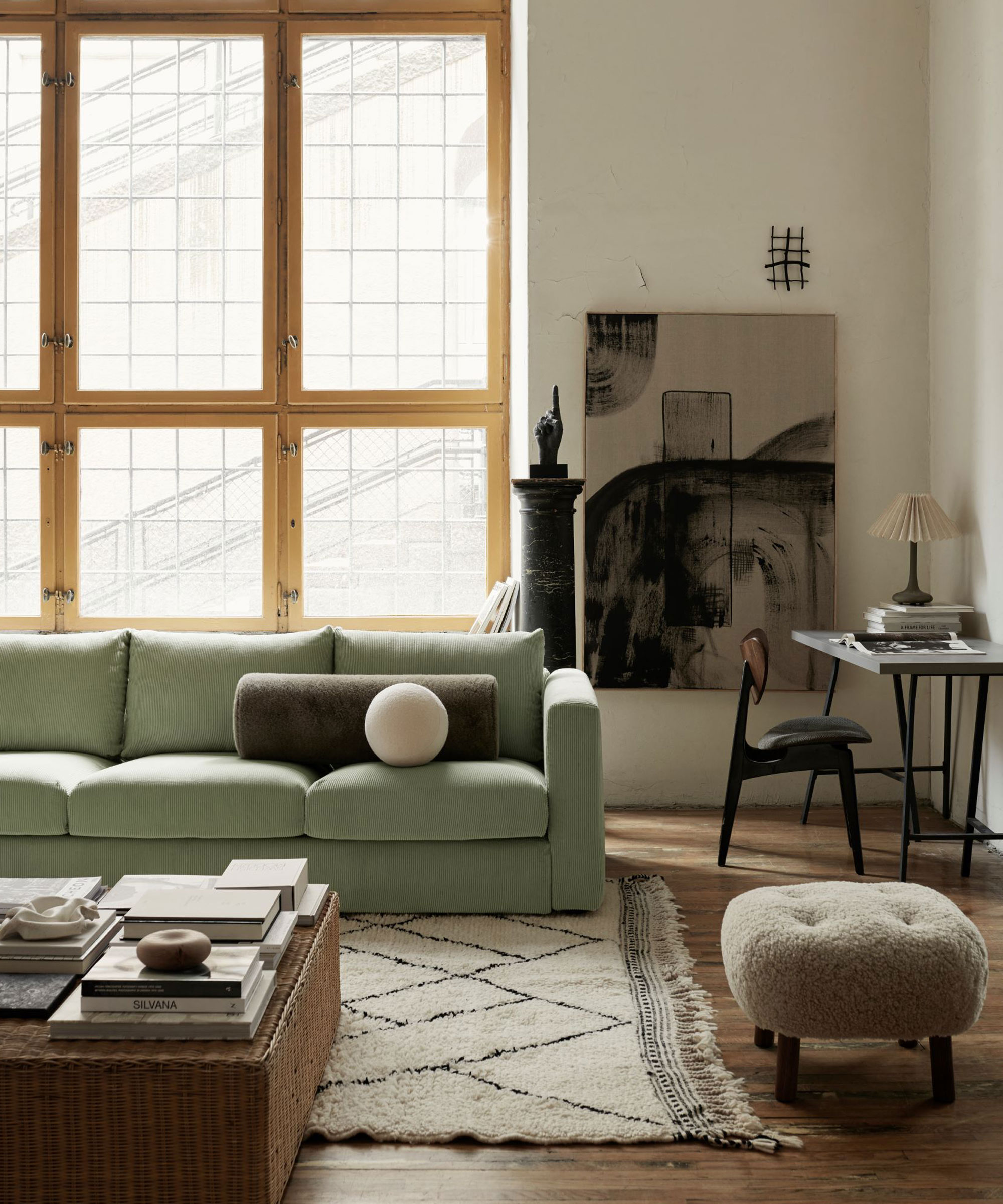 Modern living room with green sofa, dark wood flooring, cream rug, woven wicker style coffee table, upholstered ottoman with dark wood legs, large window with wooden frames, desk area with black chair, black desk, table lamp