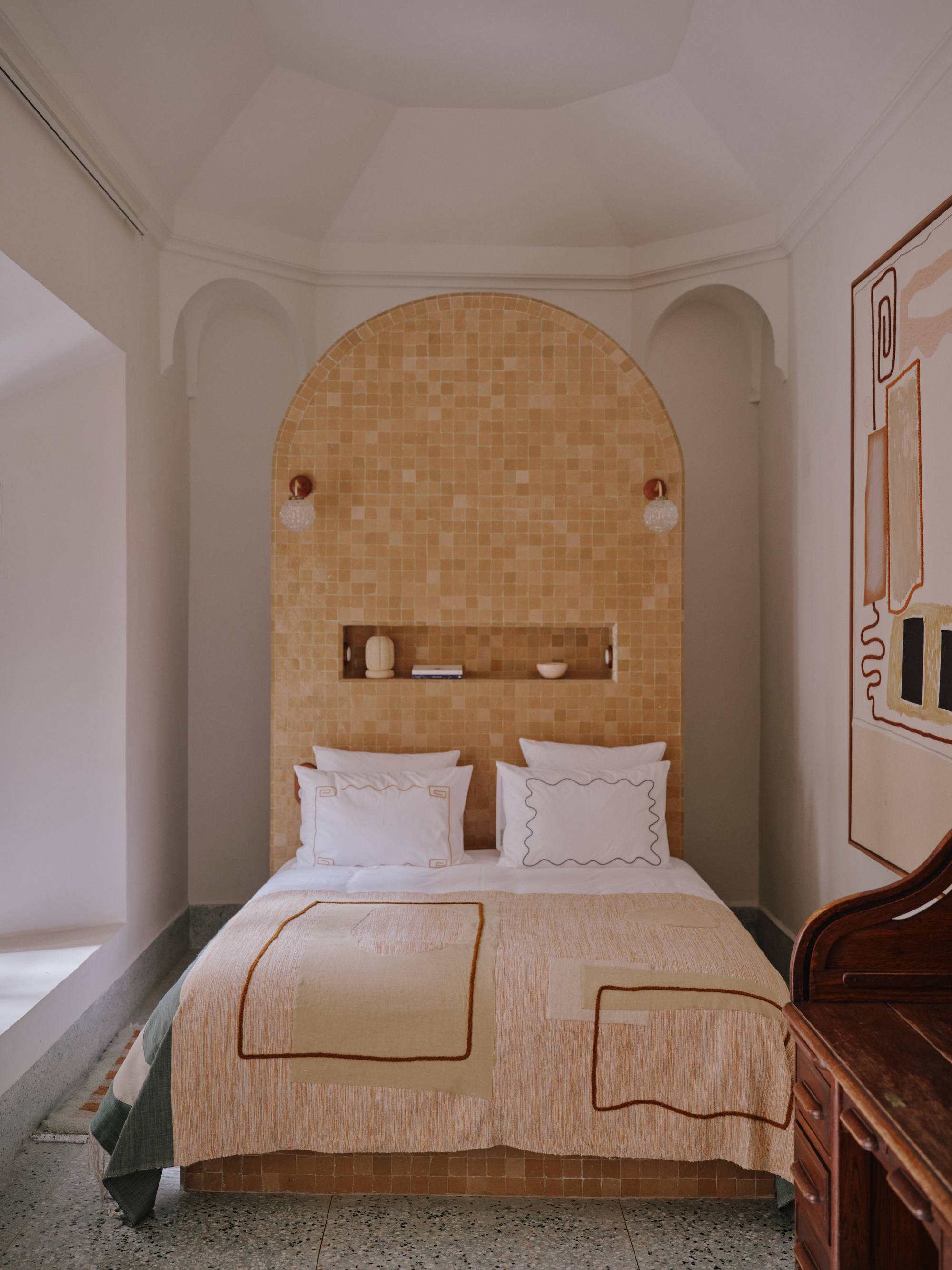 Image of a white room with an orange bed in the middle. The headboard is oversized and completely covered in orange mosaic tiles.