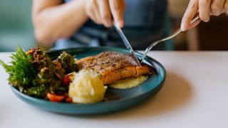 Woman eating salmon salad with knife and fork, grilled salmon, salad and potato on a plate