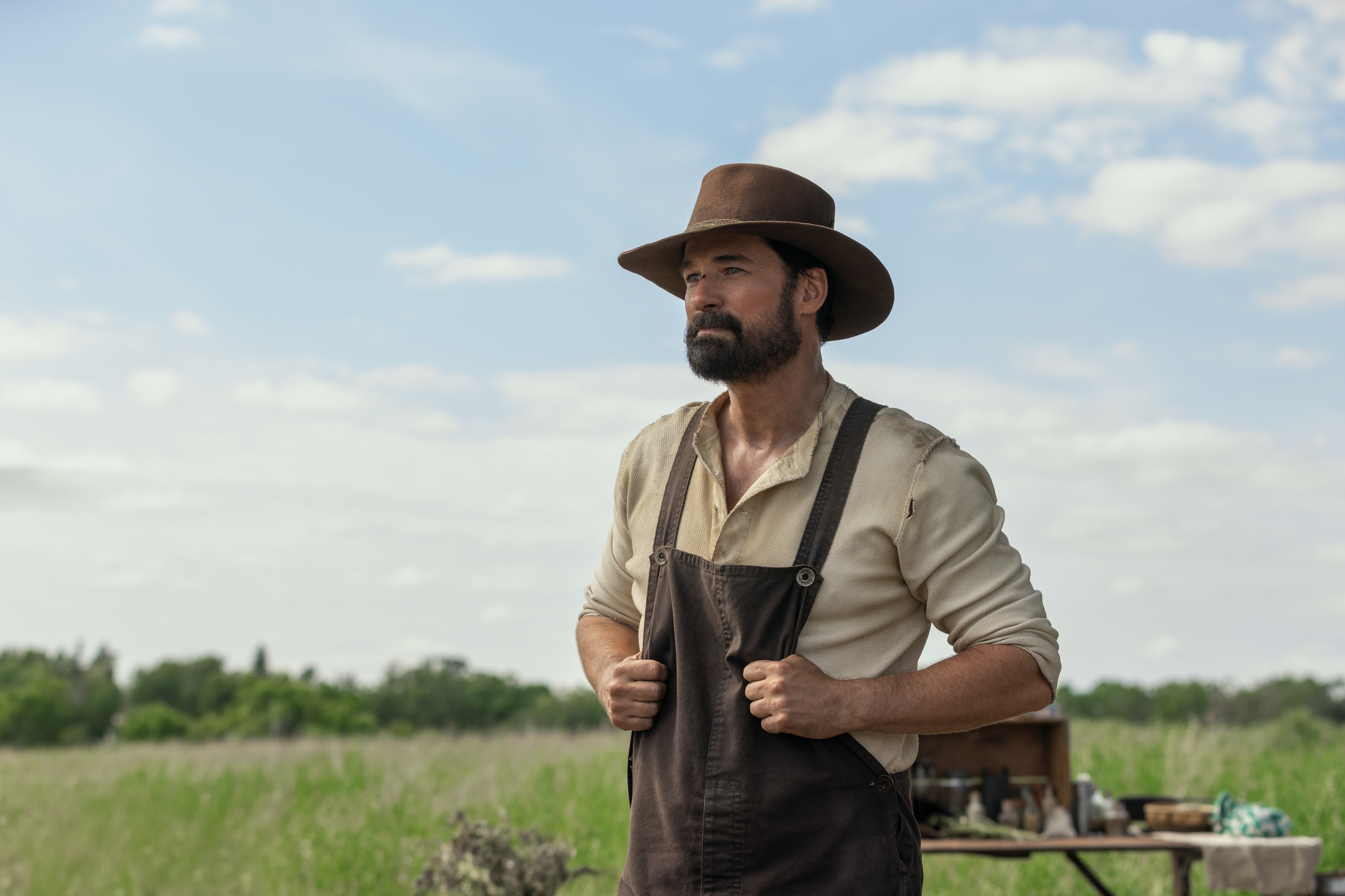 Actor Warren Christie as John Edwards wears 19th-century clothing with a hat and overalls while standing outdoors in front of a work station, in Netflix's 'Little House on the Prairie.'