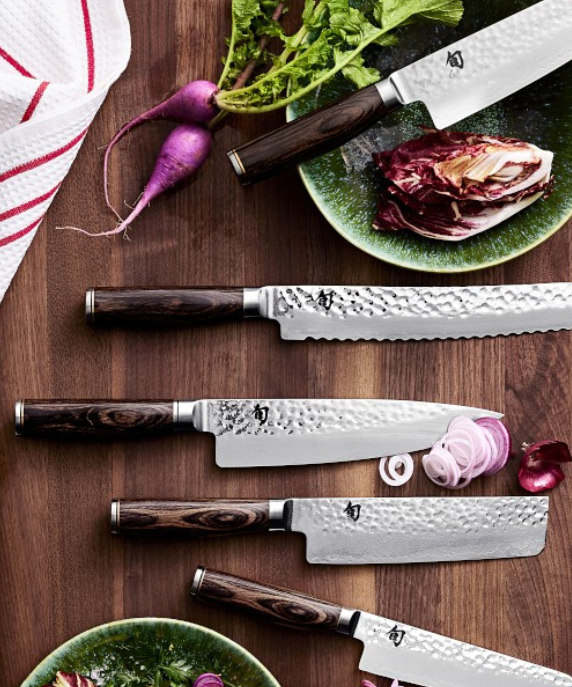A selection of Shun knoves on a dark wood cutting board, surrounded by fresh veg