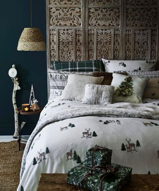 A rustic Christmas-themed bedroom with ornate headboard, dark teal wall paint decor, woven lampshade, branch decor with white and green festive motif bedding