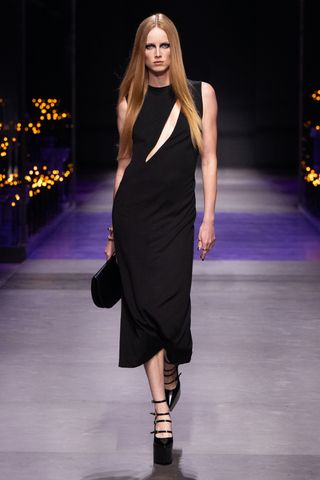 A female model wearing a long black sleeveless dress and black platform shoes walking down a runway.