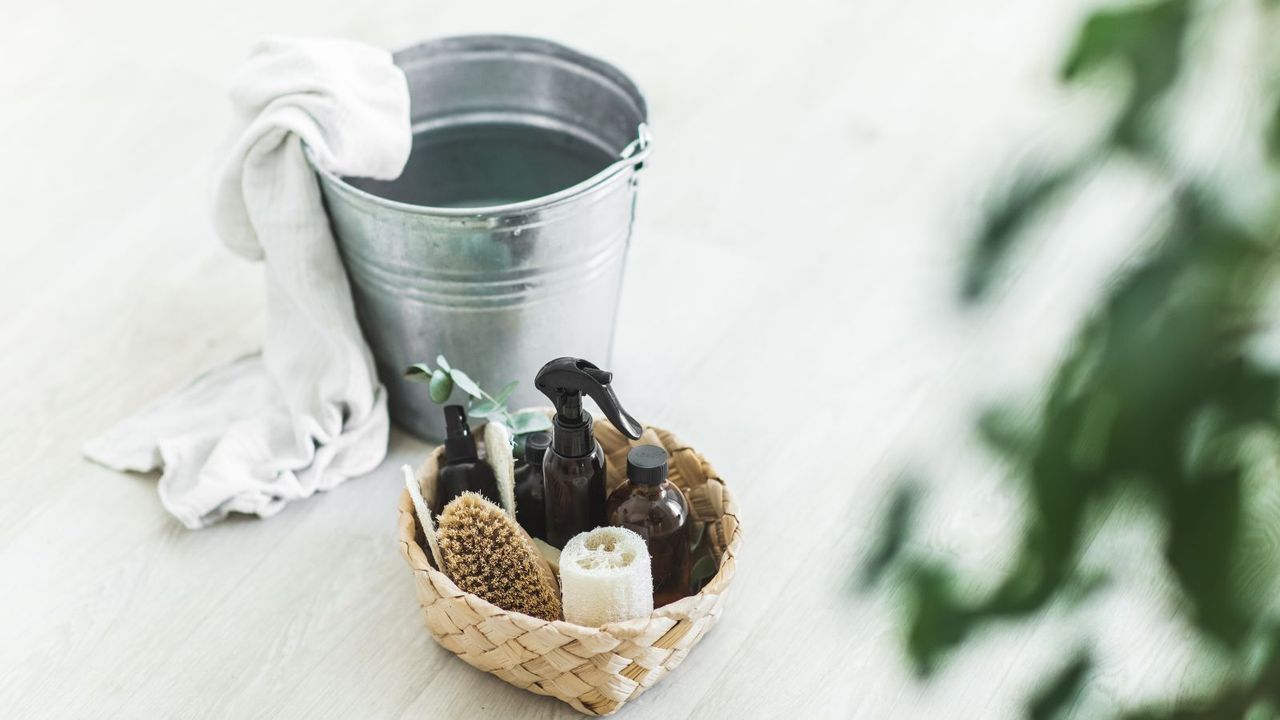 A silver mop bucket on the floor next to a wicker basket of amber cleaning bottles