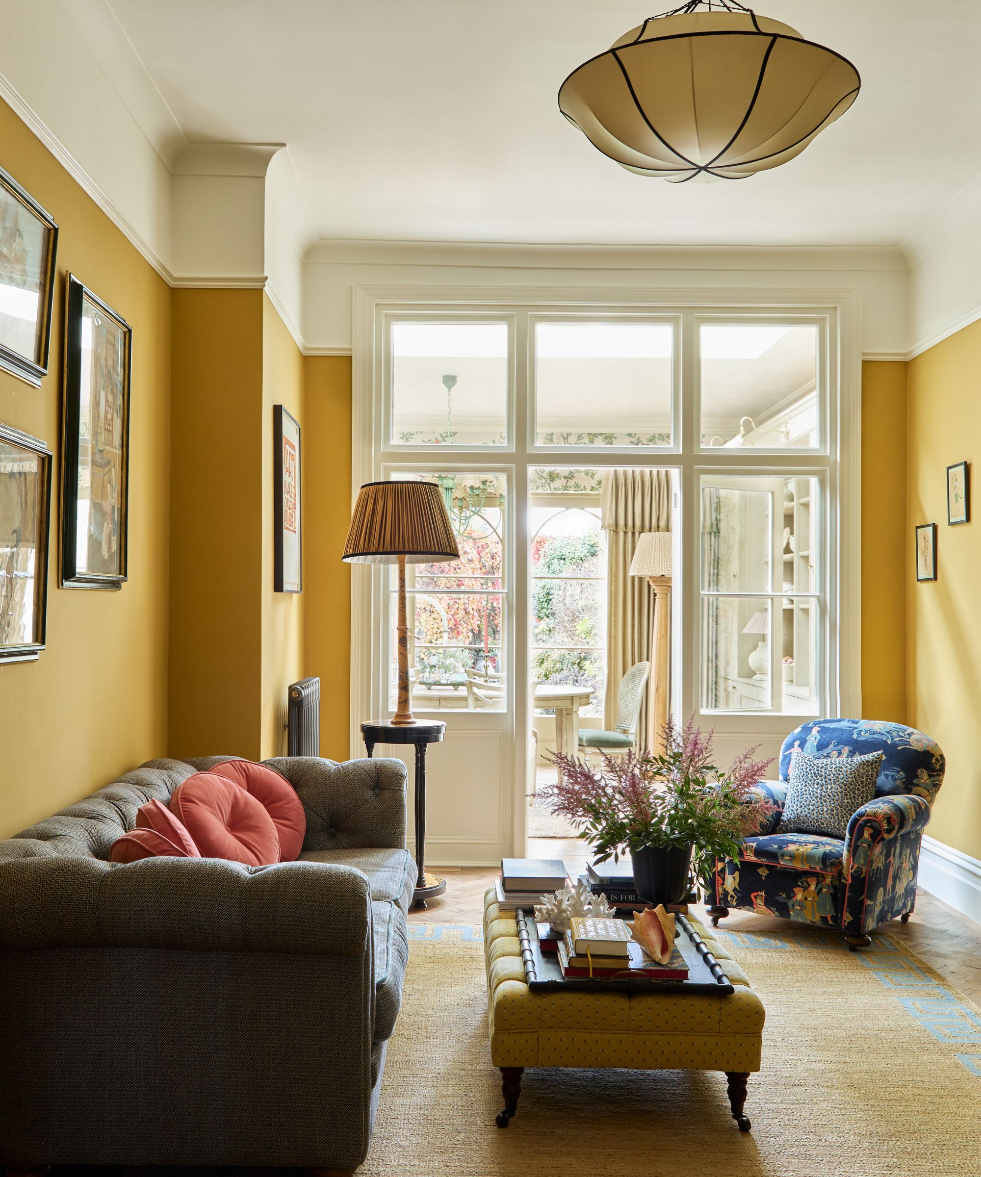 Living room with India Yellow walls, glass doors looking into a conservatory, a chesterfield grey sofa, a yellow upholstered ottoman with a tray of decor on top, and a floral armchair in the corner.