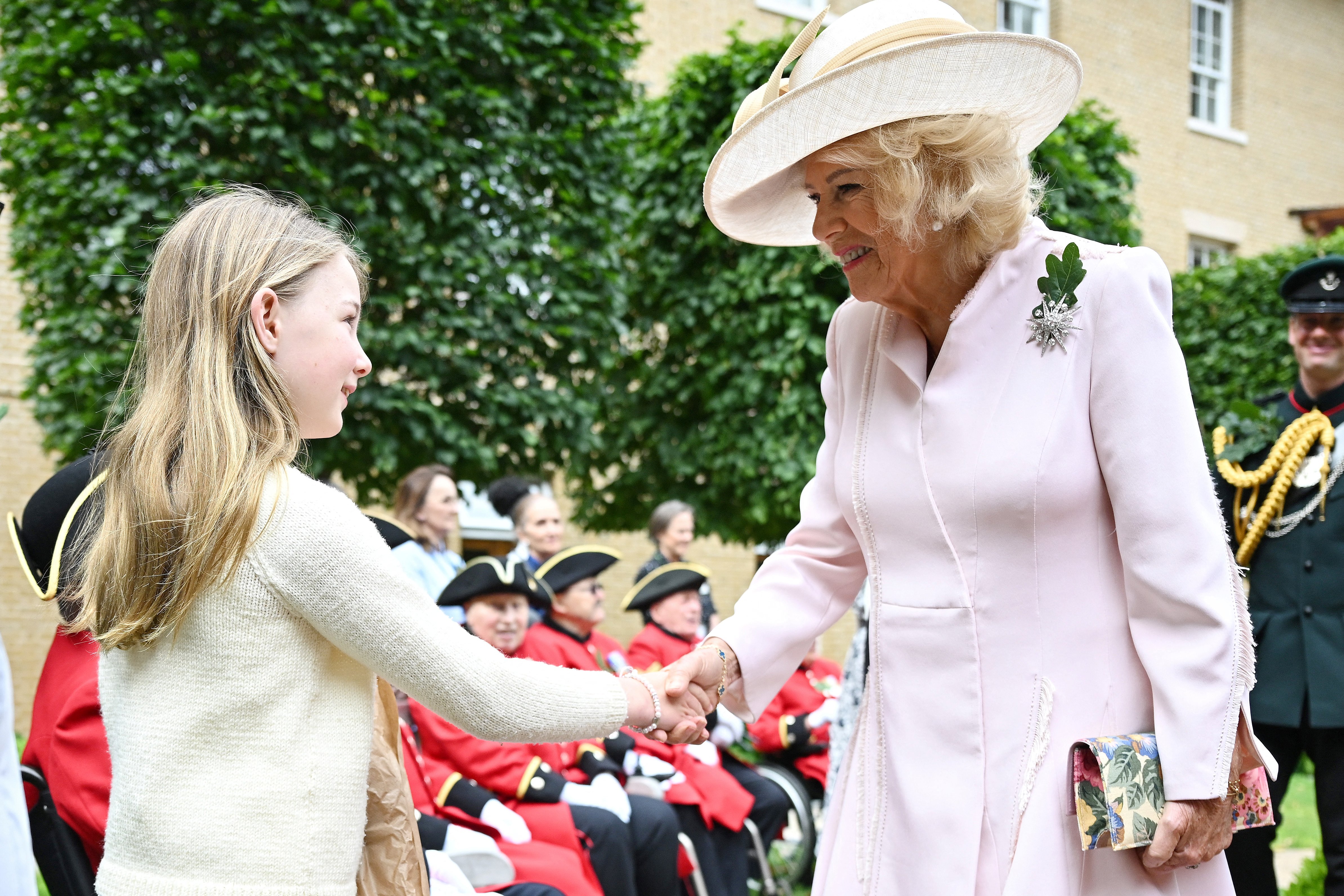 Queen Camilla wearing a pink coat shaking hands with a girl