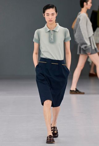 The polo shirt trend on the runway on a model wearing a gray polo with navy long shorts and fisherman sandals.